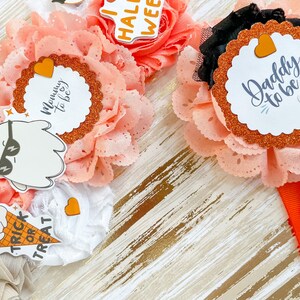 K&ouml;nnte beinhalten: Nahaufnahme von handgefertigten Baby-Haarb&auml;ndern mit Halloween-Motiv. Die Haarb&auml;nder haben pfirsichfarbene Stoffblumen, Glitzerakzente und dekorative Aufkleber mit Aufschriften wie "Mommy to be" und "Daddy to be".