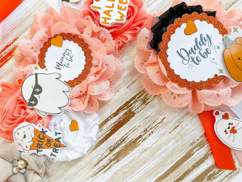 K&ouml;nnte beinhalten: Nahaufnahme von handgefertigten Baby-Haarb&auml;ndern mit Halloween-Motiv. Die Haarb&auml;nder haben pfirsichfarbene Stoffblumen, Glitzerakzente und dekorative Aufkleber mit Aufschriften wie "Mommy to be" und "Daddy to be".