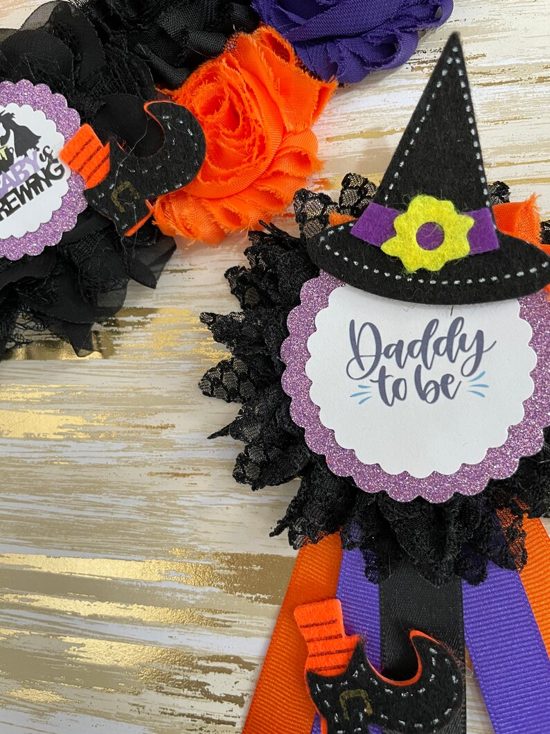 Puede incluir: Favor de fiesta de Halloween con un sombrero de bruja negro, cinta morada y negra y un c&iacute;rculo de purpurina morado con el texto "Daddy to be".