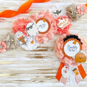 K&ouml;nnte beinhalten: Halloween-Babyparty-Dekorationen mit floralen Arrangements mit Papierschnitten von Geistern, K&uuml;rbissen und den Texten "Mommy to be" und "Daddy to be". Die Farbpalette umfasst Pfirsich, Orange und Wei&szlig;.