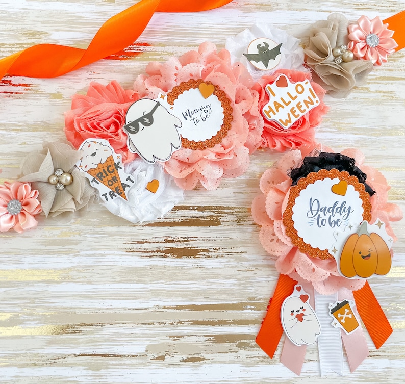 K&ouml;nnte beinhalten: Halloween-Babyparty-Dekorationen mit floralen Arrangements mit Papierschnitten von Geistern, K&uuml;rbissen und den Texten "Mommy to be" und "Daddy to be". Die Farbpalette umfasst Pfirsich, Orange und Wei&szlig;.