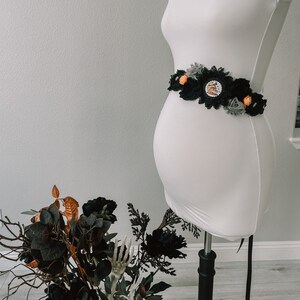 May include: A black and gray maternity sash with orange and white flowers and a black lace trim. The sash is tied around a white maternity dress on a mannequin.