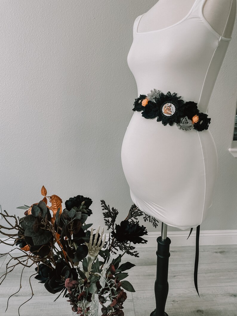 May include: A black and gray maternity sash with orange and white flowers and a black lace trim. The sash is tied around a white maternity dress on a mannequin.