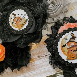 May include: Two black and orange Halloween themed baby shower sashes. One sash says "A baby is brewing" and features a witch in a cauldron. The other sash says "Daddy to be" and features a witch in a cauldron with a bat.