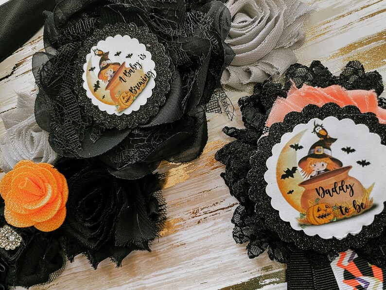May include: Two black and orange Halloween themed baby shower sashes. One sash says "A baby is brewing" and features a witch in a cauldron. The other sash says "Daddy to be" and features a witch in a cauldron with a bat.