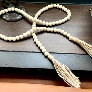 May include: A long strand of natural wood beads with two tassels made of natural fiber. The beads are white and the tassels are a light beige color.