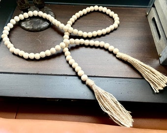May include: A long strand of natural wood beads with two tassels made of natural fiber. The beads are white and the tassels are a light beige color.