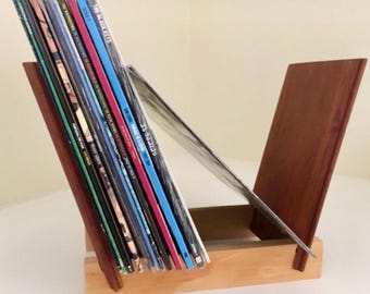 May include: Wooden vinyl record holder with a dark brown angled support and a natural wood base. The holder displays a collection of vinyl records with colorful album covers. The design is simple and functional, perfect for music enthusiasts.