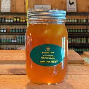 May include: A glass jar filled with peach jam. The jar has a silver lid and a green and white label that reads "Peach Jam" and includes the name and address of Twin Oak Farms. The jar is sitting on a wooden surface.