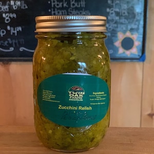 May include: A glass jar filled with green zucchini relish. The jar has a green label with the text "Twin Oak Farms Stockton, ND" and "Zucchini Relish" printed on it. The label also lists the ingredients: Zucchini, Red & Green Peppers, Onions, Sugar, Vinegar, Salt & Spices.