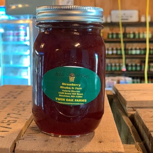 May include: A glass jar filled with red strawberry rhubarb jam. The jar has a green label with the text "Strawberry Rhubarb Jam" and the name of the farm, "Twin Oak Farms".