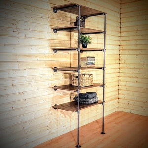 May include: A six-shelf industrial pipe shelving unit with wooden shelves. The shelves are filled with various items, including a potted plant, books, a woven basket, and folded towels.