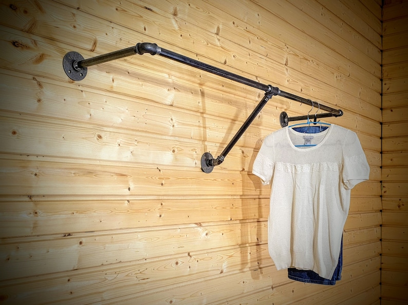 Puede incluir: Un perchero de ropa de tuber&iacute;a industrial negra montado en una pared de madera clara. El perchero sostiene una camisa blanca de manga corta y unos vaqueros azules. El perchero est&aacute; hecho de tuber&iacute;as y accesorios negros.