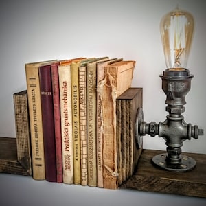 May include: A set of rustic wooden bookends with a vintage industrial pipe lamp. The lamp has a clear glass bulb and a black metal base. The bookends are holding a collection of old books with titles in Latvian.