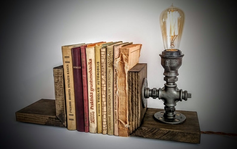 May include: A set of rustic wooden bookends with a vintage industrial pipe lamp. The lamp has a clear glass bulb and a black metal base. The bookends are holding a collection of old books with titles in Latvian.