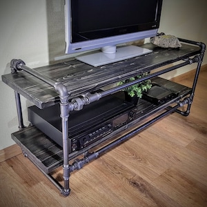 May include: A black metal TV stand with two shelves. The stand is made of industrial pipes and has a rustic wood top. A flat screen TV is sitting on the top shelf.