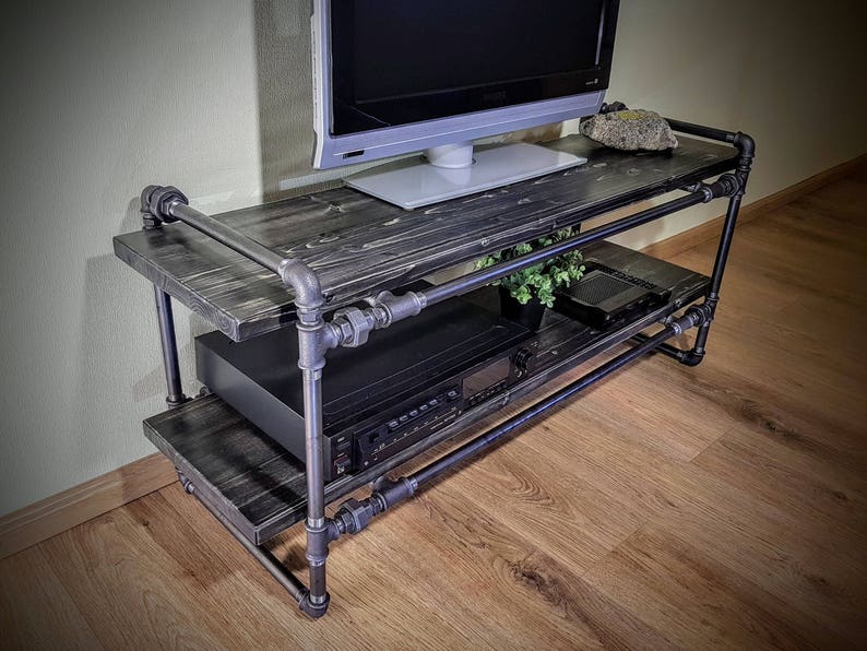May include: A black metal TV stand with two shelves. The stand is made of industrial pipes and has a rustic wood top. A flat screen TV is sitting on the top shelf.