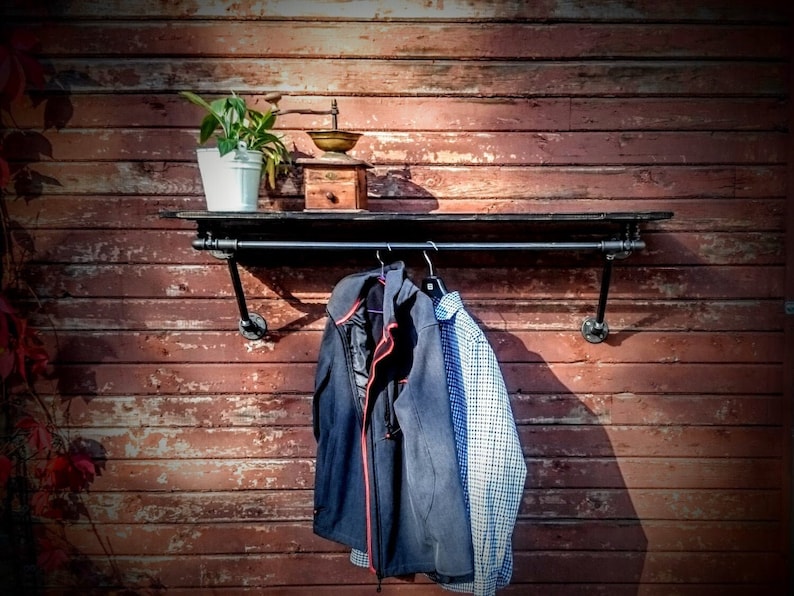 May include: A black metal shelf with a pipe design, mounted on a red wooden wall. The shelf has a black metal rod for hanging items. Two jackets and a shirt are hanging on the rod. A small potted plant and an antique coffee grinder are on the shelf.