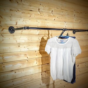Puede incluir: Un perchero de ropa de tuber&iacute;a industrial negra montado en una pared de tablones de madera clara. Una camisa blanca de manga corta y unos vaqueros azules cuelgan del perchero. El perchero est&aacute; hecho de tuber&iacute;as y accesorios negros.
