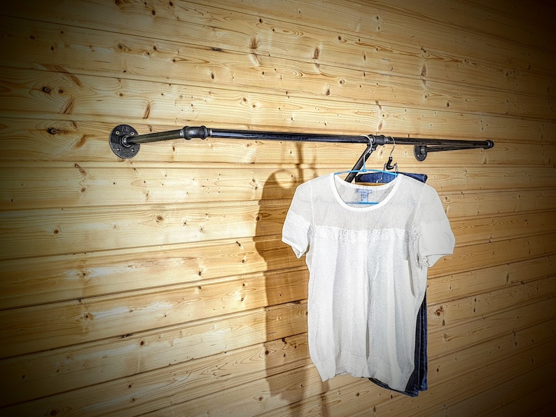 Puede incluir: Un perchero de ropa de tuber&iacute;a industrial negra montado en una pared de tablones de madera clara. Una camisa blanca de manga corta y unos vaqueros azules cuelgan del perchero. El perchero est&aacute; hecho de tuber&iacute;as y accesorios negros.