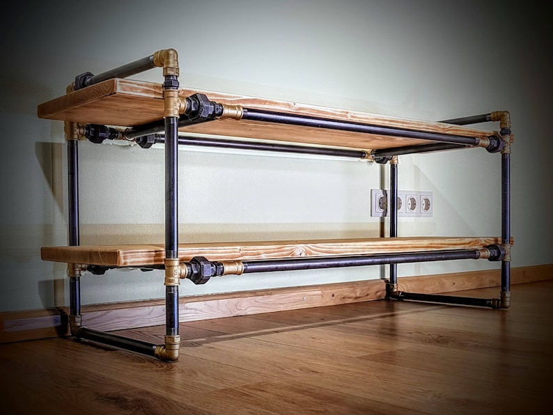 May include: A two-tiered shelf unit made of black metal pipes and wooden shelves. The pipes have gold-colored fittings. The shelf unit is standing on a wooden floor.