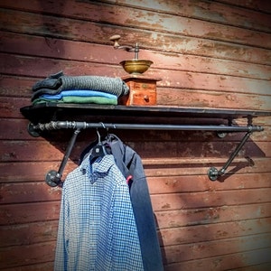 May include: A black metal pipe shelf with a black metal rod for hanging clothes. The shelf is mounted on a red wooden wall. A stack of folded clothing and a vintage coffee grinder are on the shelf. A blue and white plaid shirt is hanging on the rod.