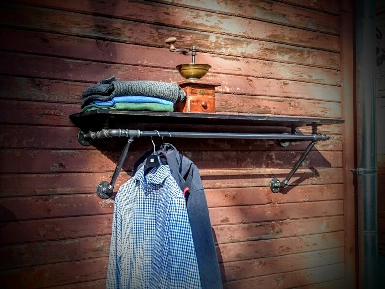 May include: A black metal pipe shelf with a black metal rod for hanging clothes. The shelf is mounted on a red wooden wall. A stack of folded clothing and a vintage coffee grinder are on the shelf. A blue and white plaid shirt is hanging on the rod.
