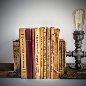 May include: A rustic wooden bookend set with a vintage industrial pipe lamp. The bookends are made of dark wood and have a natural finish. The lamp is made of black metal pipe and has a clear glass bulb. The lamp is lit and casting a warm glow. The bookends are holding a collection of vintage books with titles in different languages.