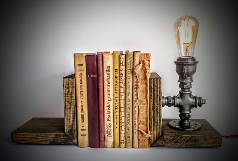 May include: A rustic wooden bookend set with a vintage industrial pipe lamp. The bookends are made of dark wood and have a natural finish. The lamp is made of black metal pipe and has a clear glass bulb. The lamp is lit and casting a warm glow. The bookends are holding a collection of vintage books with titles in different languages.