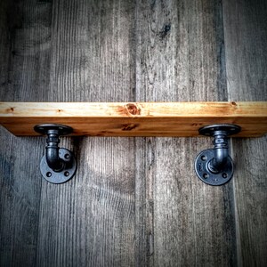 May include: A wooden shelf with black metal pipe brackets. The shelf is mounted on a wood wall.