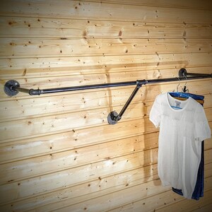 Puede incluir: Un perchero de ropa de tuber&iacute;a industrial negra montado en una pared de madera clara. Cuelgan una camisa blanca de manga corta y unos vaqueros azules. El perchero est&aacute; hecho de tuber&iacute;as y accesorios de metal negro.
