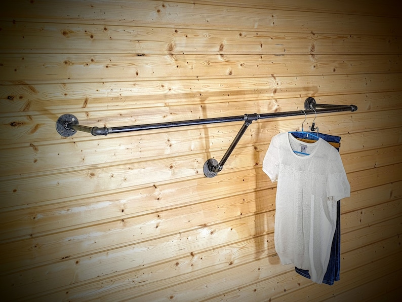 Puede incluir: Un perchero de ropa de tuber&iacute;a industrial negra montado en una pared de madera clara. Cuelgan una camisa blanca de manga corta y unos vaqueros azules. El perchero est&aacute; hecho de tuber&iacute;as y accesorios de metal negro.