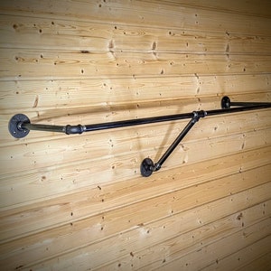 Puede incluir: Un perchero de ropa de tuber&iacute;a industrial negra montado en una pared de madera marr&oacute;n claro. El perchero tiene una barra horizontal con una extensi&oacute;n en &aacute;ngulo recto, soportada por tuber&iacute;as anguladas y bridas circulares.