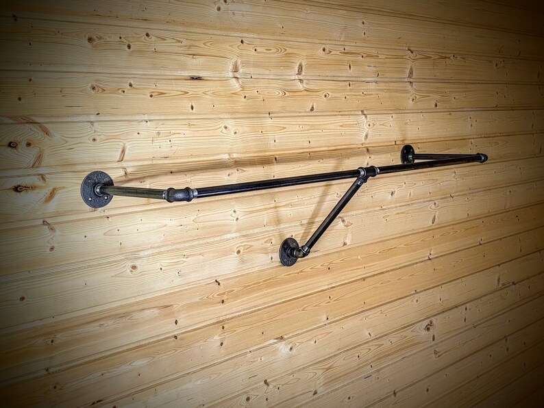 Puede incluir: Un perchero de ropa de tuber&iacute;a industrial negra montado en una pared de madera marr&oacute;n claro. El perchero tiene una barra horizontal con una extensi&oacute;n en &aacute;ngulo recto, soportada por tuber&iacute;as anguladas y bridas circulares.