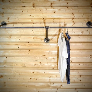 Puede incluir: Un perchero de ropa de tuber&iacute;a industrial negra montado en una pared de madera clara. El perchero sostiene una parte superior blanca y pantalones azul oscuro en perchas. El perchero est&aacute; hecho de tuber&iacute;as y accesorios negros.
