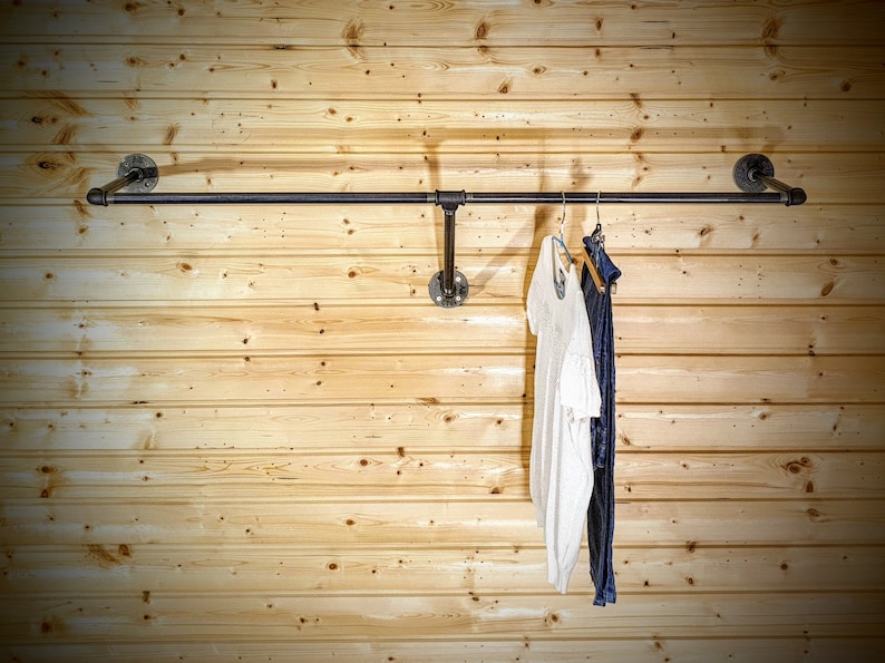 Puede incluir: Un perchero de ropa de tuber&iacute;a industrial negra montado en una pared de madera clara. El perchero sostiene una parte superior blanca y pantalones azul oscuro en perchas. El perchero est&aacute; hecho de tuber&iacute;as y accesorios negros.