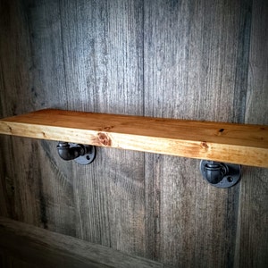 May include: A wooden shelf with black metal pipe brackets. The shelf is mounted on a wood wall.