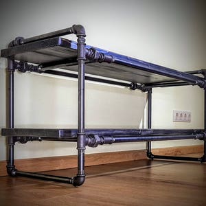 May include: A black metal shelf unit with two tiers. The shelves are made of wood and are supported by black metal pipes. The shelf unit is standing on a wooden floor.