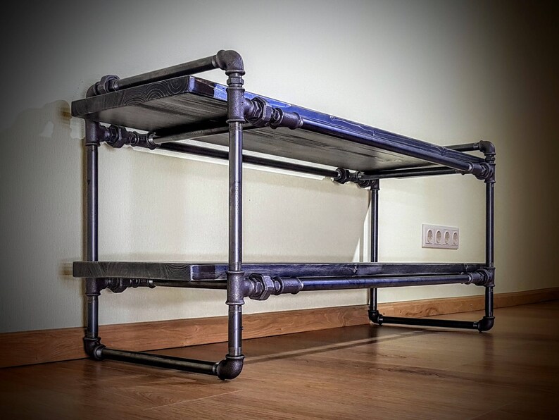 May include: A black metal shelf unit with two tiers. The shelves are made of wood and are supported by black metal pipes. The shelf unit is standing on a wooden floor.