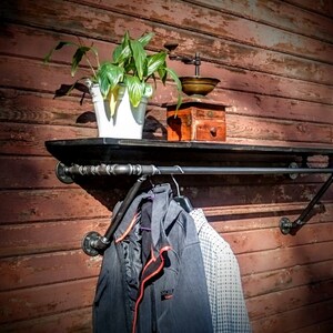 May include: A black metal pipe shelf and clothes rack mounted on a red wooden wall. The shelf has a potted plant and an antique coffee grinder on it. Two jackets hang on the rack.
