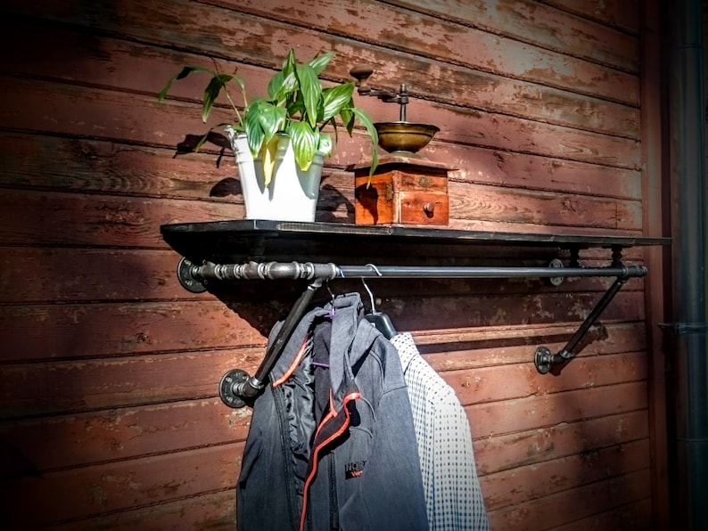 May include: A black metal pipe shelf and clothes rack mounted on a red wooden wall. The shelf has a potted plant and an antique coffee grinder on it. Two jackets hang on the rack.