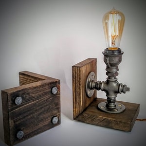 May include: A rustic wooden lamp base with a metal pipe and Edison bulb. The base is made of dark wood with a distressed finish and has a metal pipe that is attached to the base. The pipe has a metal flange and a metal bulb holder. The bulb is an Edison bulb with a clear glass and a filament that is visible when the bulb is lit.
