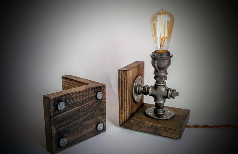 May include: A rustic wooden lamp base with a metal pipe and Edison bulb. The base is made of dark wood with a distressed finish and has a metal pipe that is attached to the base. The pipe has a metal flange and a metal bulb holder. The bulb is an Edison bulb with a clear glass and a filament that is visible when the bulb is lit.