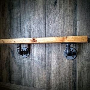 May include: A wooden shelf with black metal pipe brackets mounted on a wood wall. The shelf is made of natural wood with a light brown finish. The brackets are made of black metal and have a pipe design.
