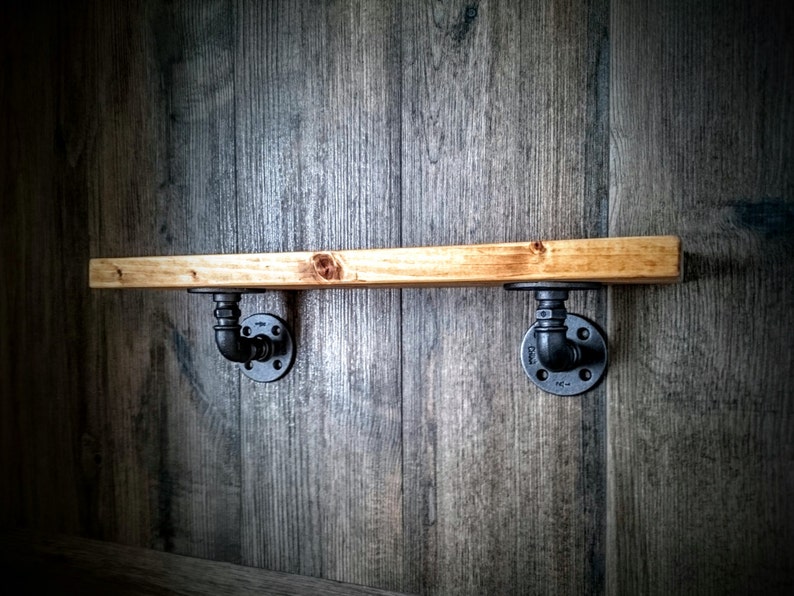 May include: A wooden shelf with black metal pipe brackets mounted on a wood wall. The shelf is made of natural wood with a light brown finish. The brackets are made of black metal and have a pipe design.