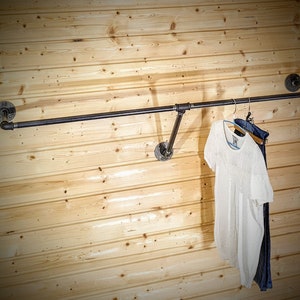 Puede incluir: Perchero de estilo industrial hecho de tuber&iacute;as de metal oscuro, montado en una pared de paneles de madera clara. Una camiseta blanca y unos vaqueros azules cuelgan del perchero. El perchero tiene una barra horizontal y soportes angulados.