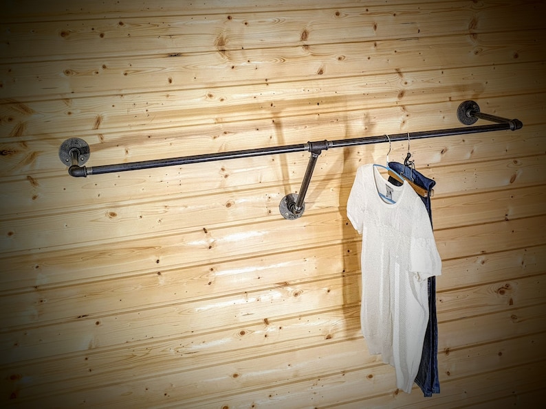Puede incluir: Perchero de estilo industrial hecho de tuber&iacute;as de metal oscuro, montado en una pared de paneles de madera clara. Una camiseta blanca y unos vaqueros azules cuelgan del perchero. El perchero tiene una barra horizontal y soportes angulados.