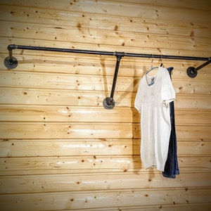 Puede incluir: Un perchero de ropa de tuber&iacute;a industrial negra montado en una pared de madera clara. Una blusa blanca y unos vaqueros azules cuelgan del perchero. El perchero est&aacute; hecho de tuber&iacute;as y accesorios negros.