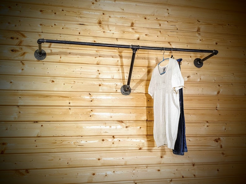 Puede incluir: Un perchero de ropa de tuber&iacute;a industrial negra montado en una pared de madera clara. Una blusa blanca y unos vaqueros azules cuelgan del perchero. El perchero est&aacute; hecho de tuber&iacute;as y accesorios negros.