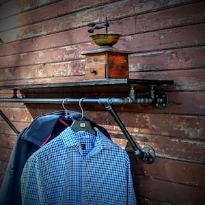 May include: A black metal pipe shelf with a black metal pipe rod attached to the bottom. The shelf is mounted to a wooden wall and has a vintage coffee grinder on top. Two shirts are hanging on the rod.